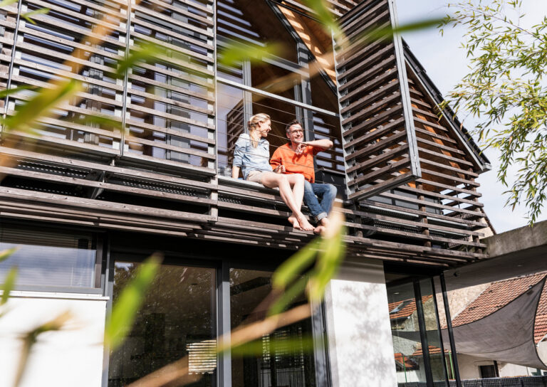 Haus in der Natur mit einem Paar, welches aus dem Balkon blickt und lächelt.
